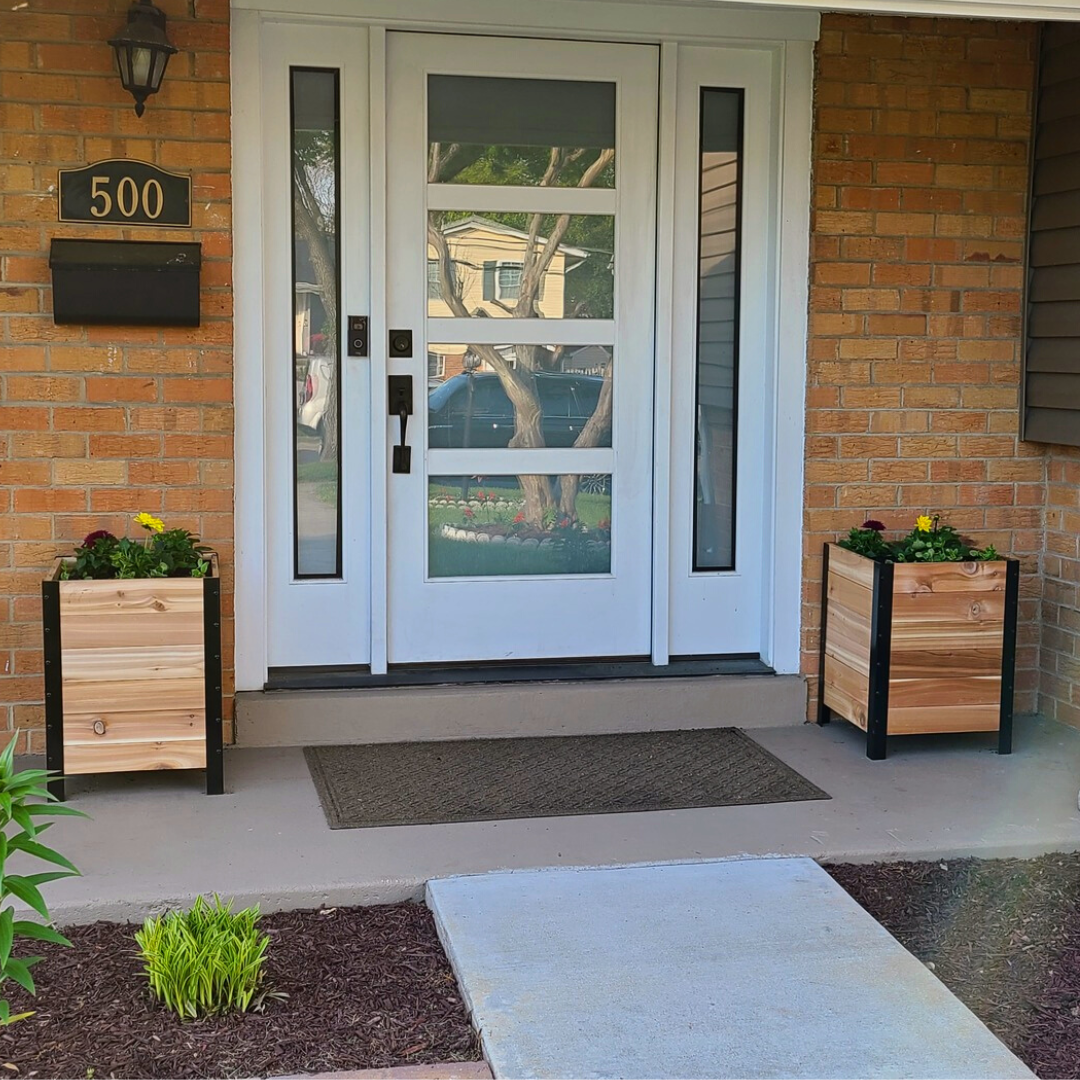 Two matching cedar planter boxes with black metal frames flank a white front door. Both planters are filled with yellow and purple flowers, sitting on a porch with red brick walls.