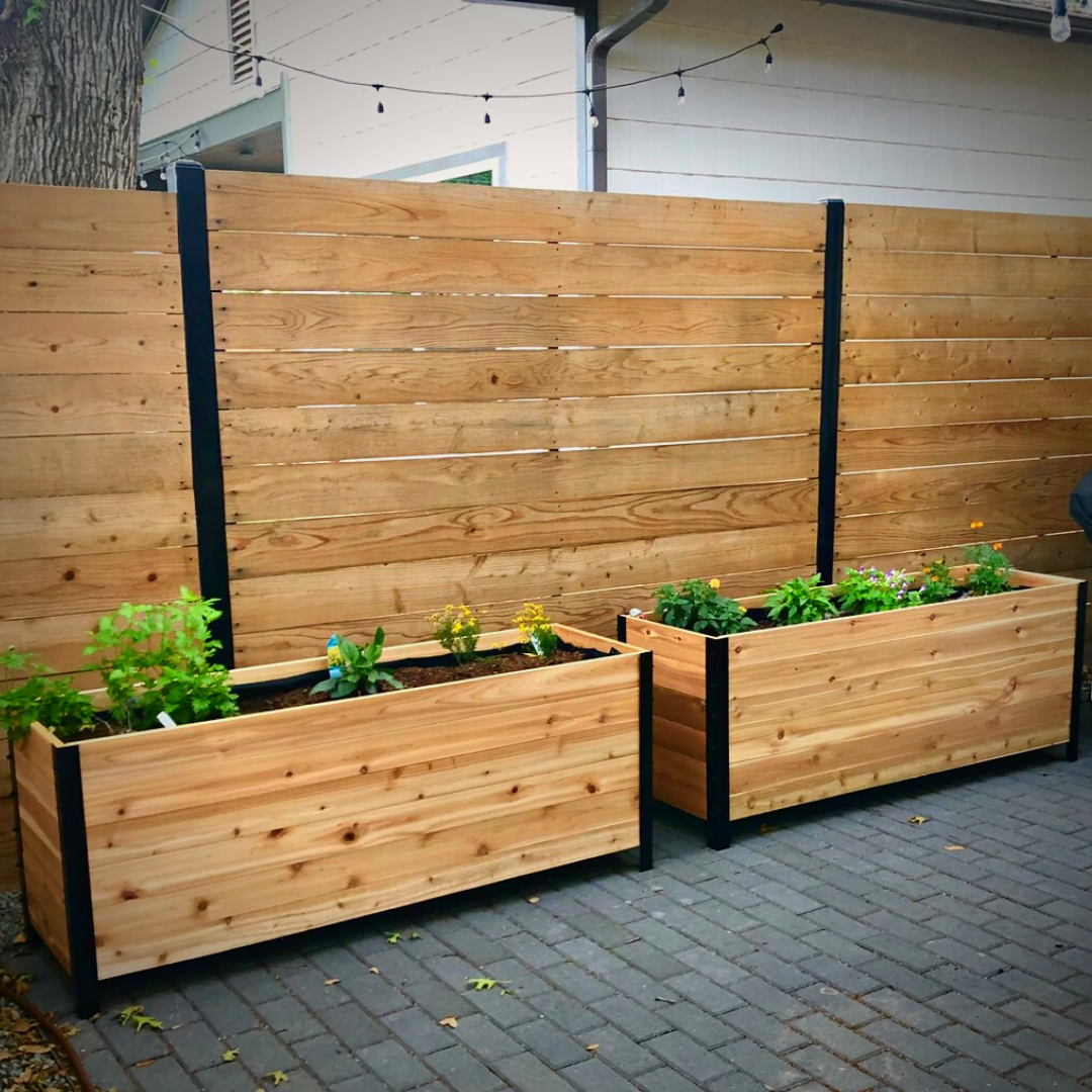 Two 18" x 60" cedar planter boxes with black steel corners placed on a patio, filled with flowers and herbs, set against a wooden privacy fence.