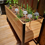 A long, raised cedar planter box with a black metal frame sits on a sunny wooden deck. It's filled with herbs marked by metal signs, including Rosemary, Thyme, Dill, and Oregano.