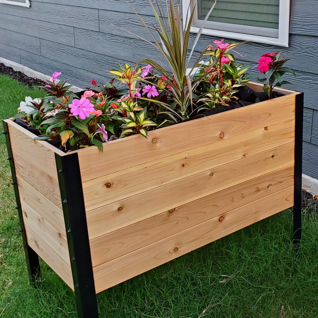 A raised cedar planter box with a black metal frame is filled with vibrant pink, white, and variegated flowers, sitting on green grass against a dark gray house wall.