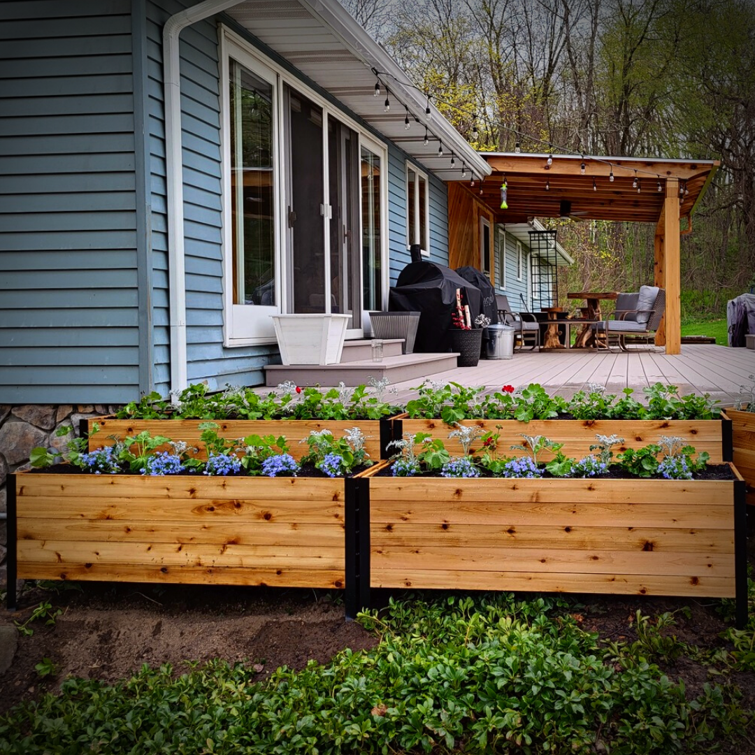Multiple 18" x 60" cedar planter boxes with black steel corners arranged in a tiered layout beside a house patio, filled with blooming flowers and greenery.