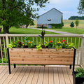 A long, raised cedar planter box with a black metal frame sits on a wooden deck railing. It is full of colorful flowers (red, yellow, orange) and mixed foliage, overlooking a grassy yard.