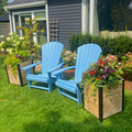 Two wooden planter boxes flank a pair of bright blue Adirondack chairs on a sunny lawn. The planters are filled with cascading red and yellow flowers and foliage, creating a charming garden seating area.