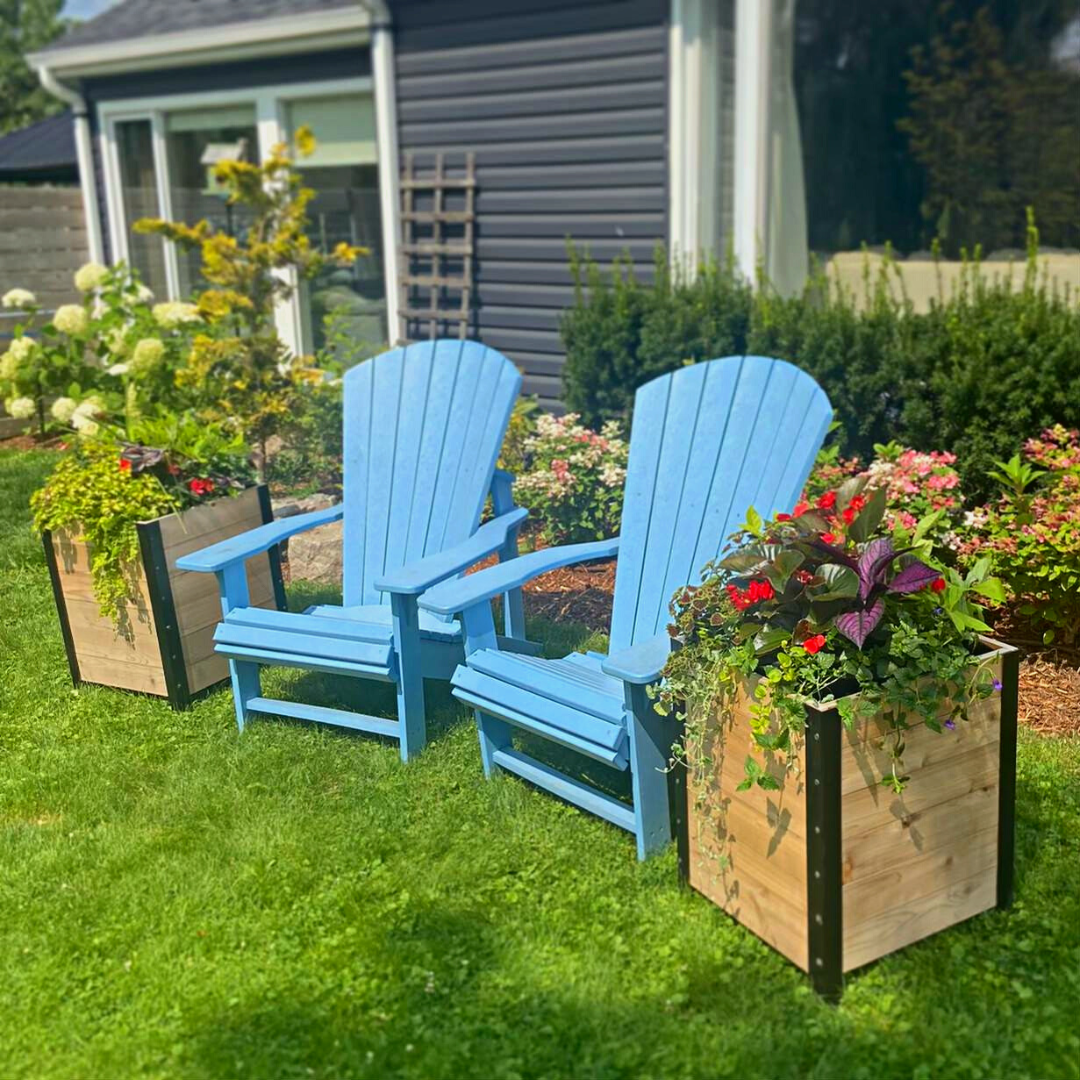 Two wooden planter boxes flank a pair of bright blue Adirondack chairs on a sunny lawn. The planters are filled with cascading red and yellow flowers and foliage, creating a charming garden seating area.