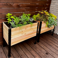 Two raised cedar planter boxes with black metal frames sit on a brown deck against a wood slat fence. The left box holds green herbs, and the right holds colorful yellow and red foliage.